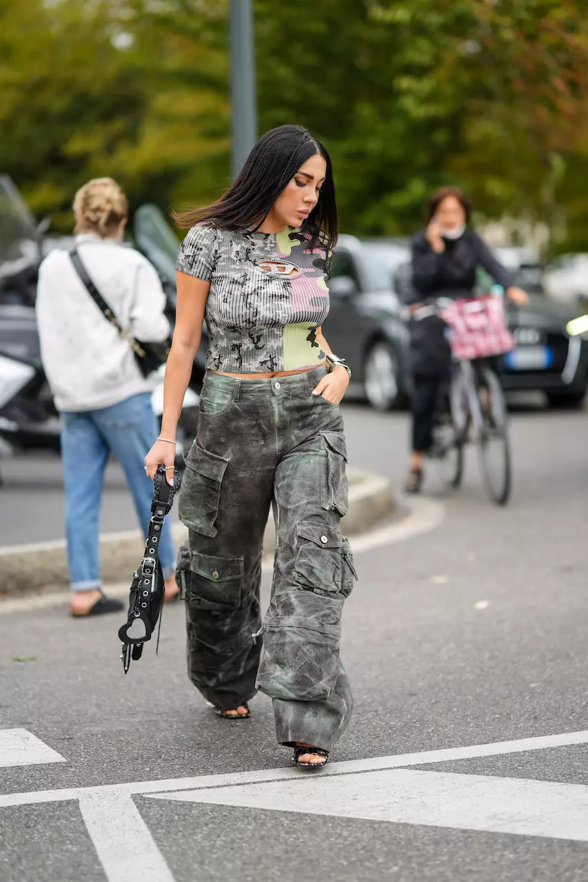 Tiffany Hsu wears a black and neon green futurist sunglasses, a white tank-top / zipper corset with suspender belt, gray faded denim large cargo pants, a green shiny leather handbag, black open toe cap plastic shoes , outside Missoni, during the Milan Fashion Week - Womenswear Spring/Summer 2023 on September 23, 2022 in Milan, Italy.
