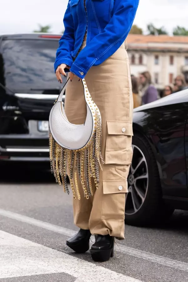 woman in blue denim asymmetric cropped jacket, leather bag with nailed studded fringed chain pendant shoulder, beige cargo pants, platform high heels shoes.